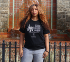 Woman wearing a black t-shirt with white 'Angry but Justified' graphic design, standing against a brick wall waterfall.
