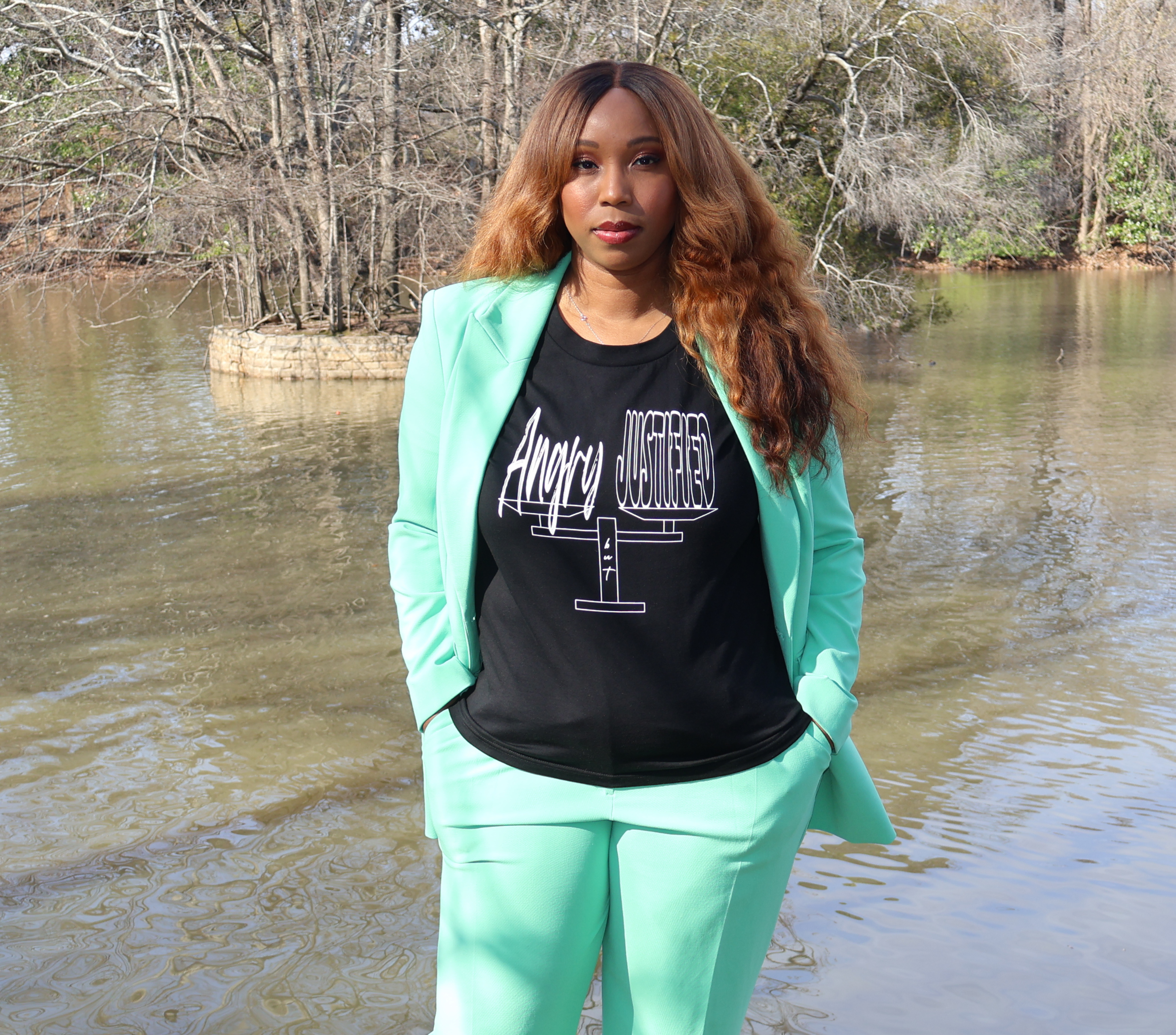 Woman wearing a green suit and black t-shirt with white 'Angry but Justified' graphic design, standing next to a lake with trees.