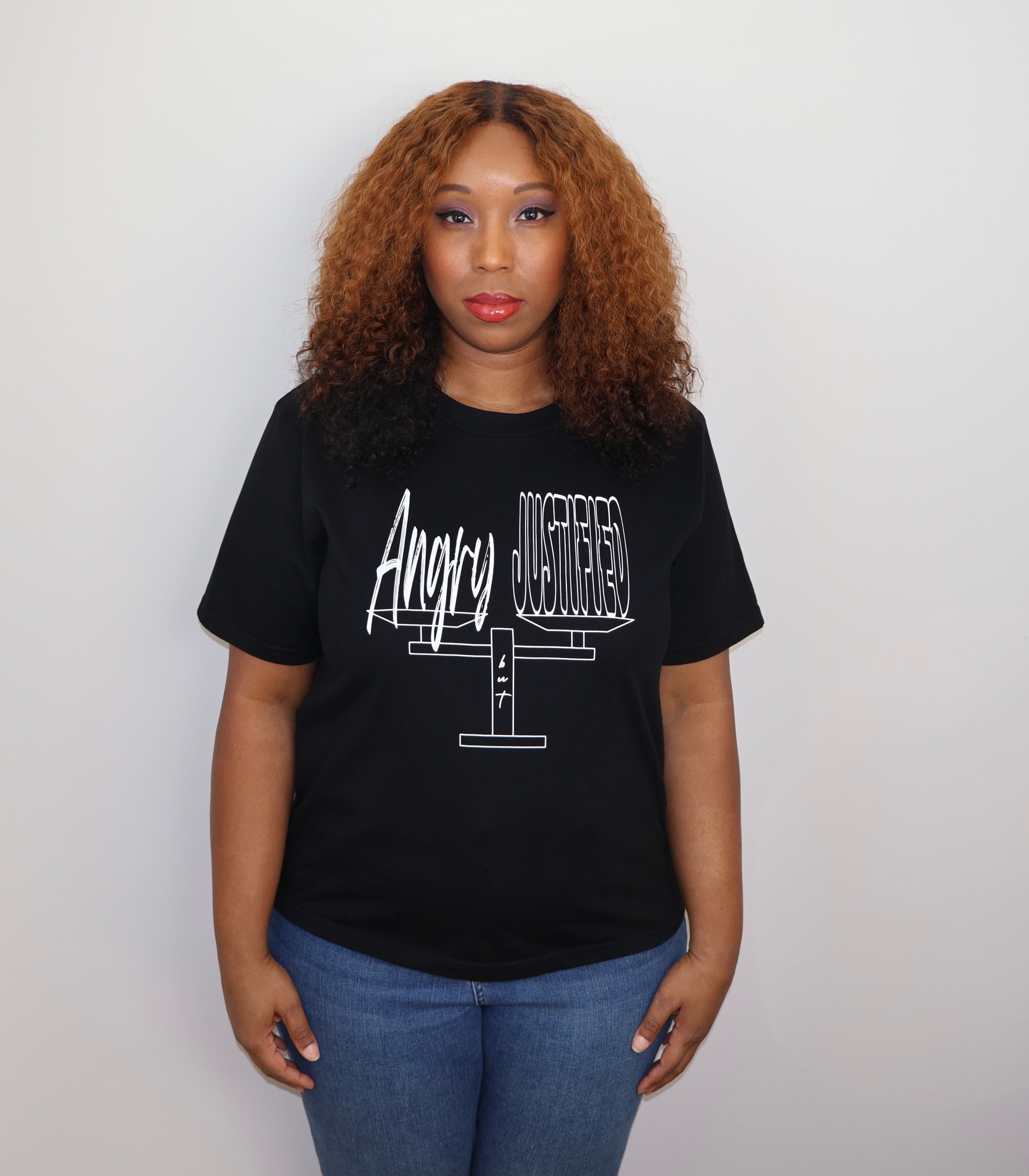 Woman wearing a black t-shirt with white 'Angry but Justified' text and graphic design, standing against a gray wall
