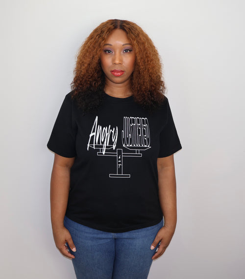 Woman wearing a black t-shirt with white 'Angry but Justified' text and graphic design, standing against a gray wall
