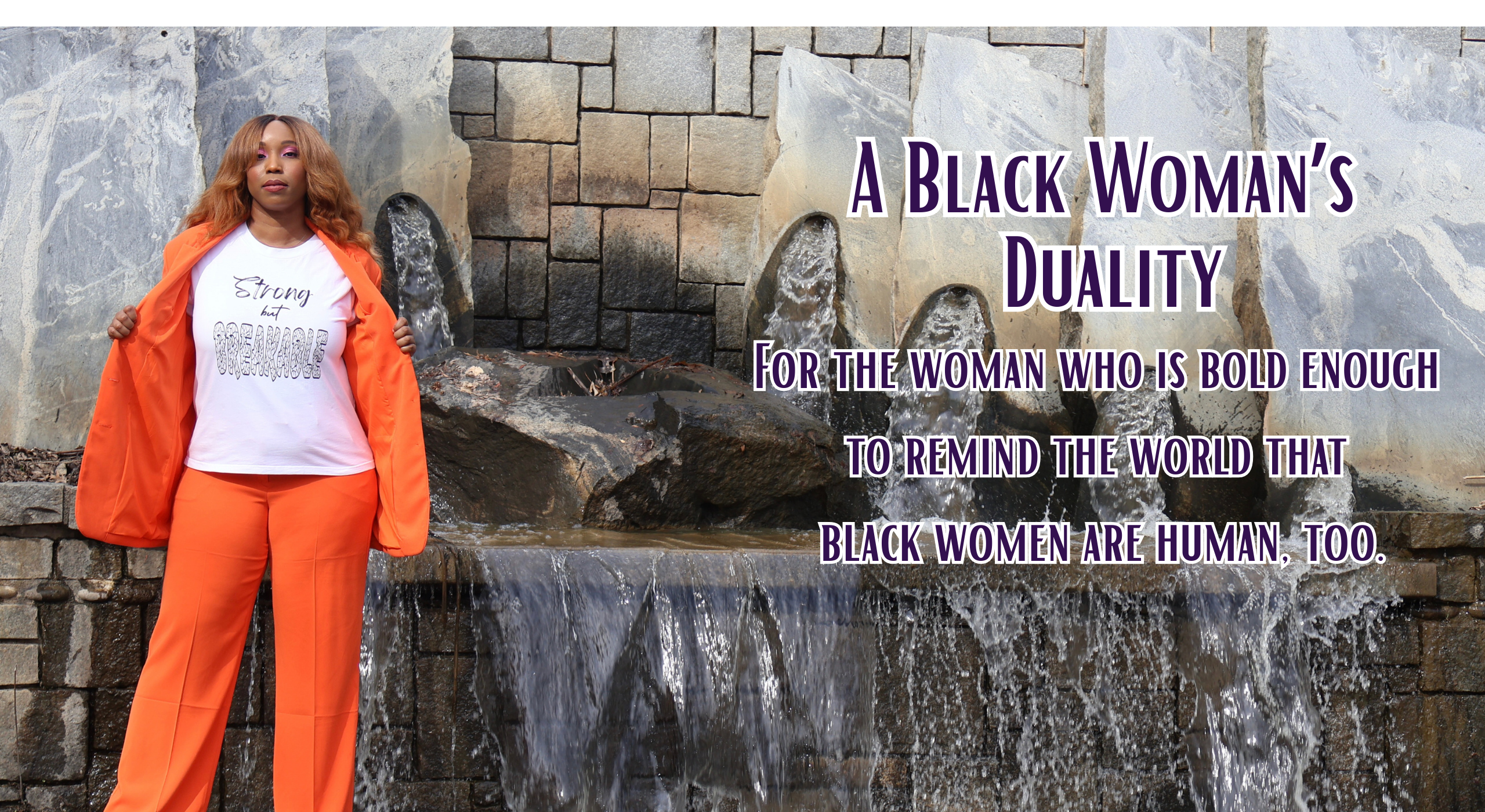 Woman wearing an orange jacket and pants with a white shirt with black 'Strong but Breakable' graphic design, standing against a stone wall waterfall.