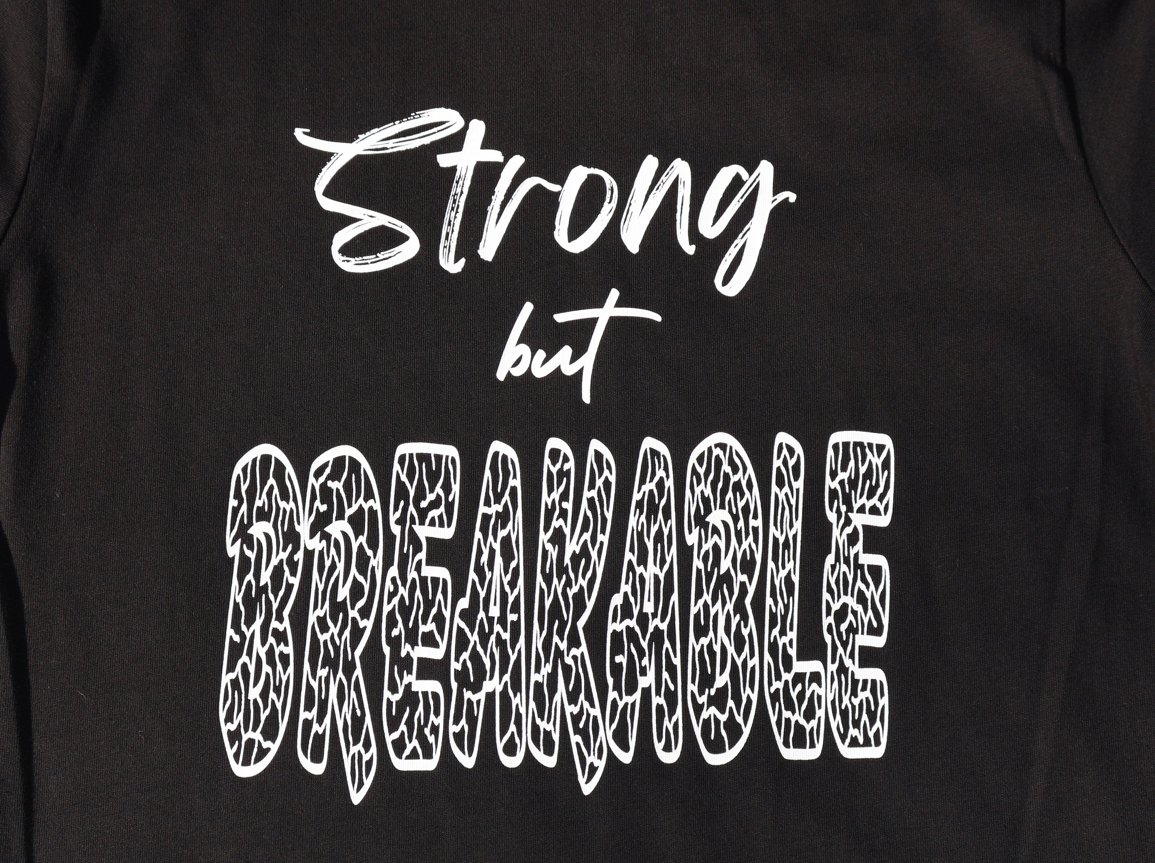 Close up of Black t-shirt with white 'Strong but Breakable' text and graphic design. 