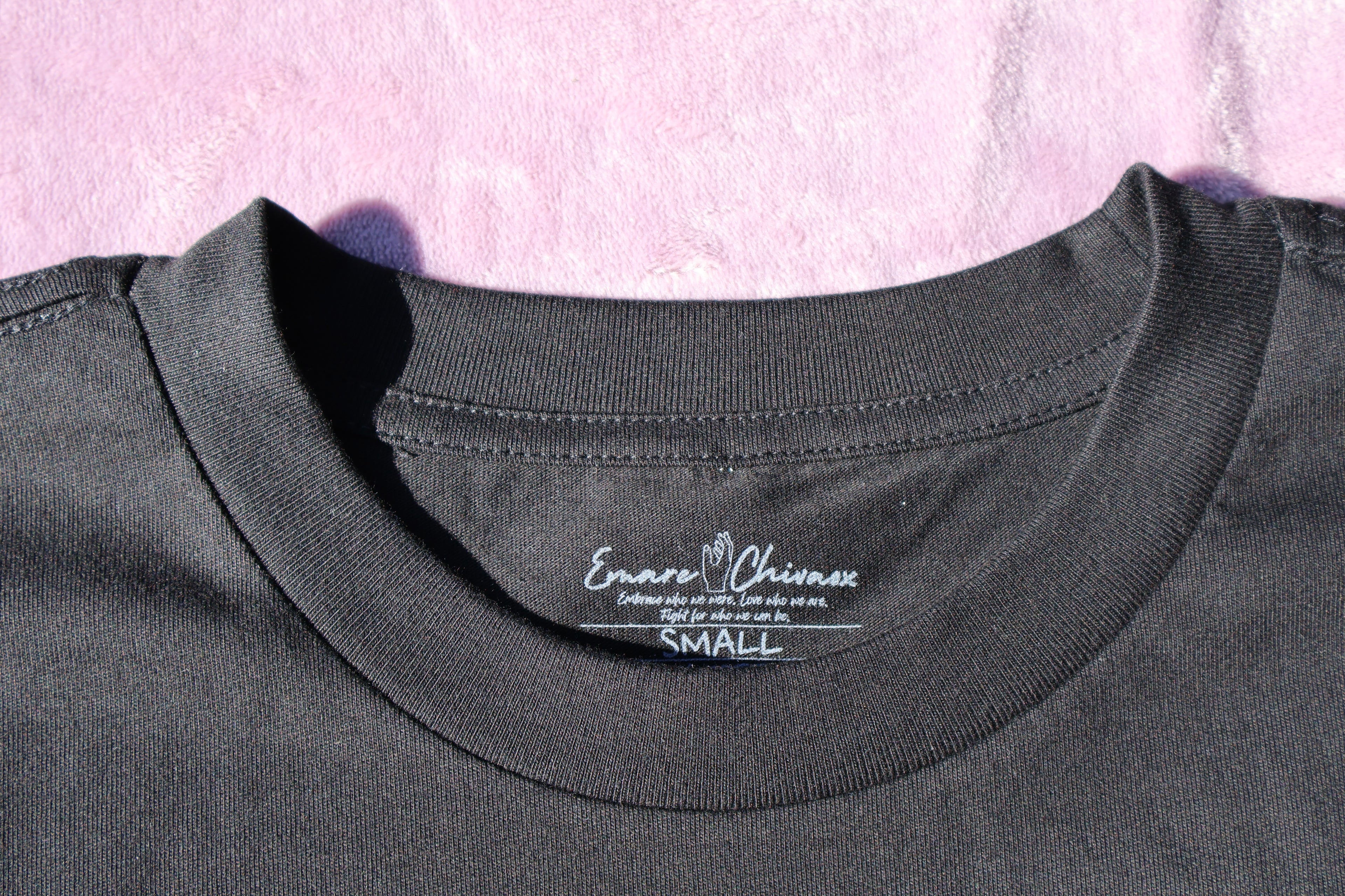 Close up of the front collar of a black t-shirt with white custom Emare Chivaox neck tag on a lavender background.