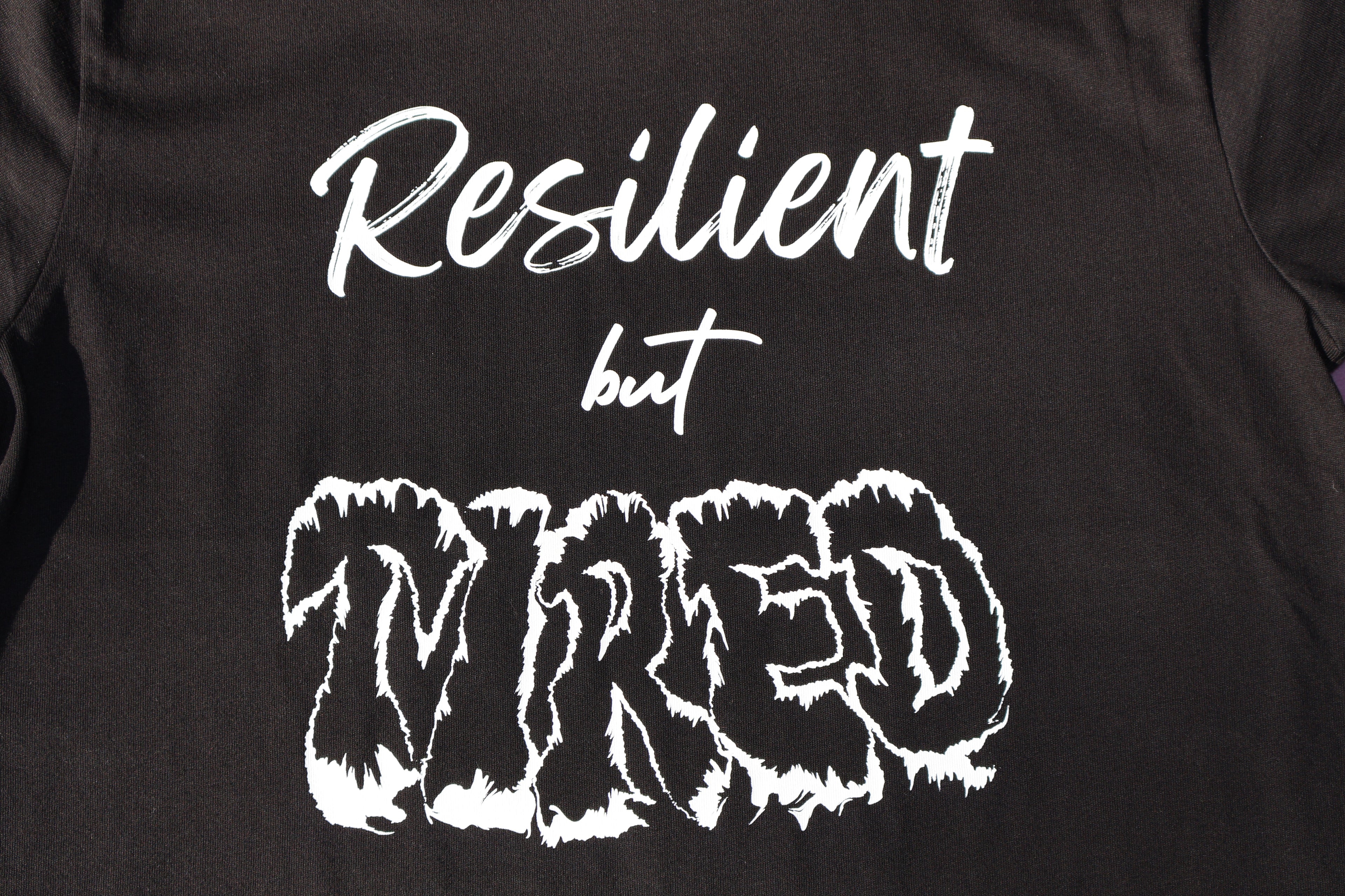Close up of Black t-shirt with white 'Resilient but Tired' text and graphic design. 