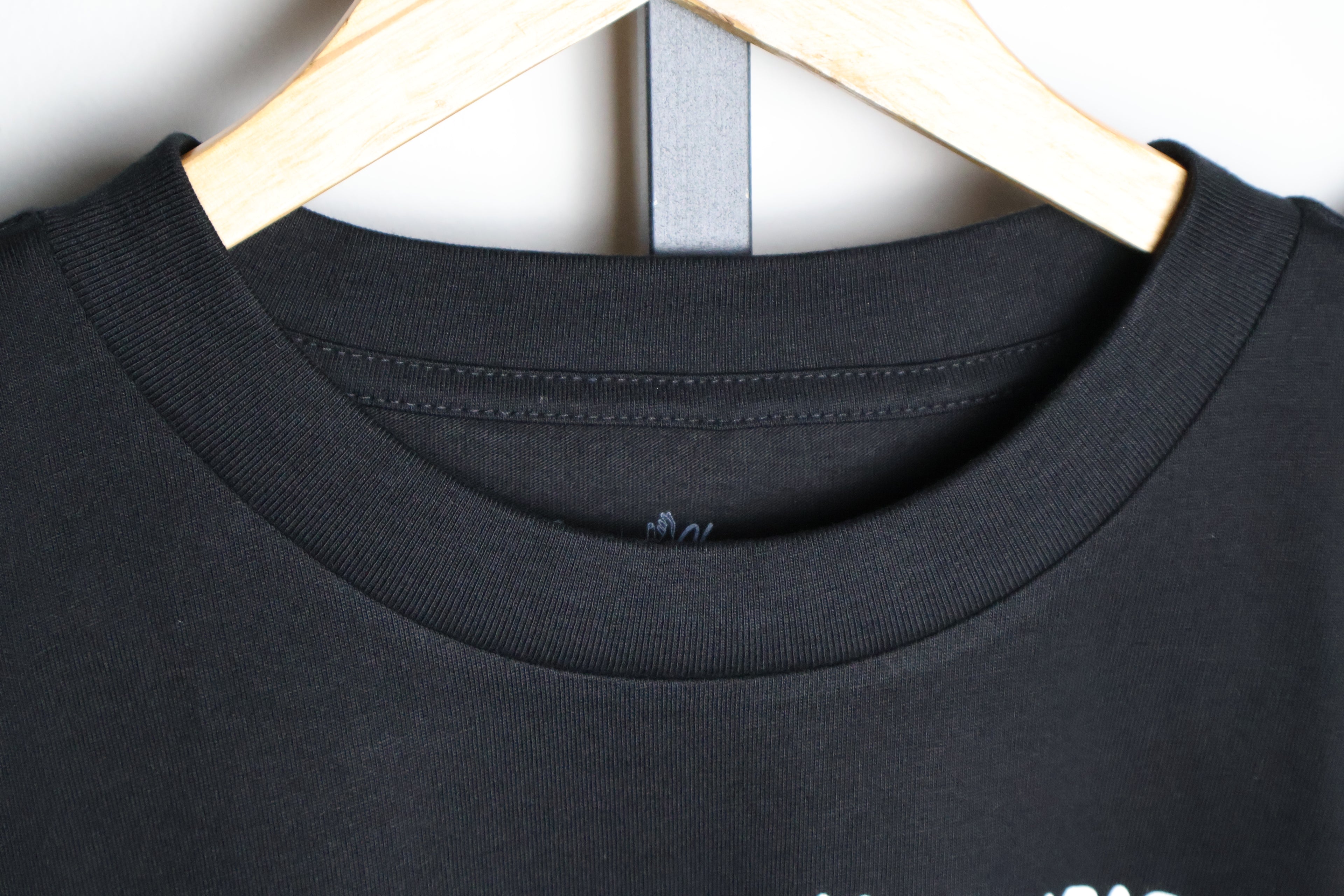Close up of black t-shirt front collar with white custom Emare Chivaox neck tag hanging on a wooden hanger.