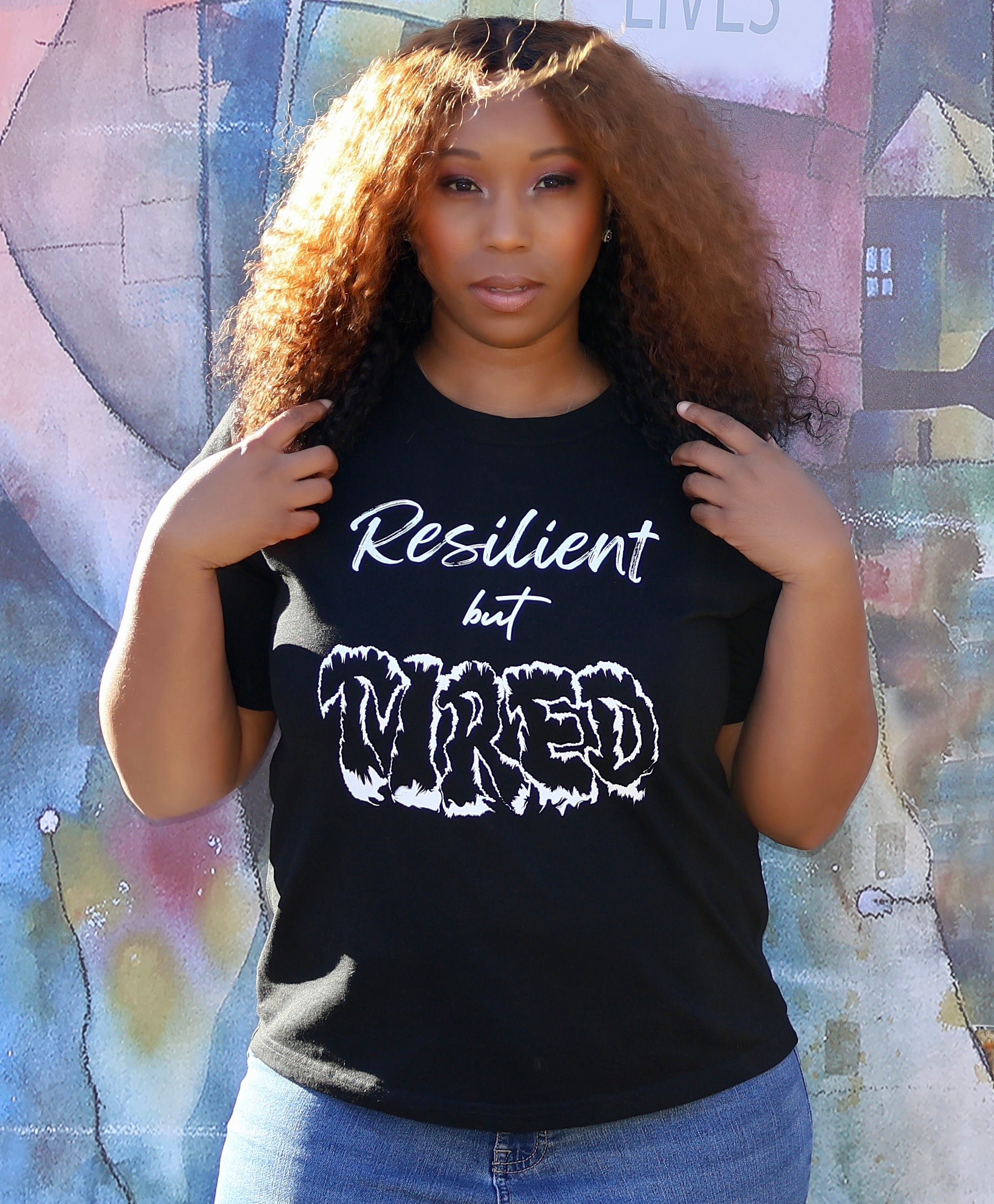 Woman wearing a black t-shirt with white 'Resilient but Tired' text and graphic design, standing in front of a colorful mural.