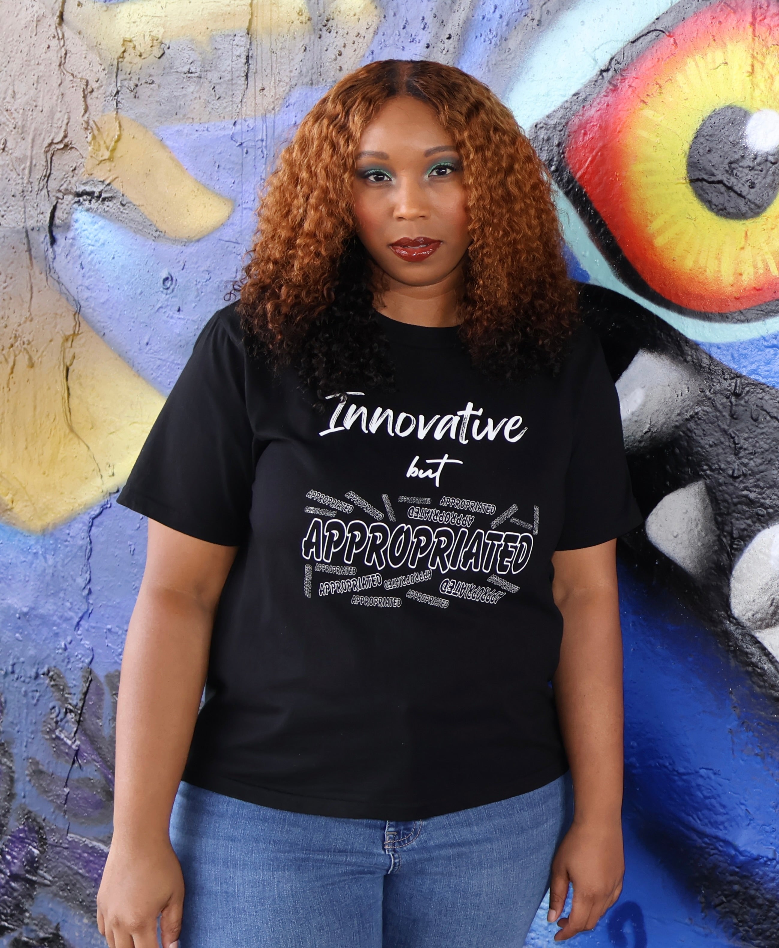 Woman wearing a black t-shirt with white 'Innovative but Appropriated' text and graphic design in front of a colorful mural.