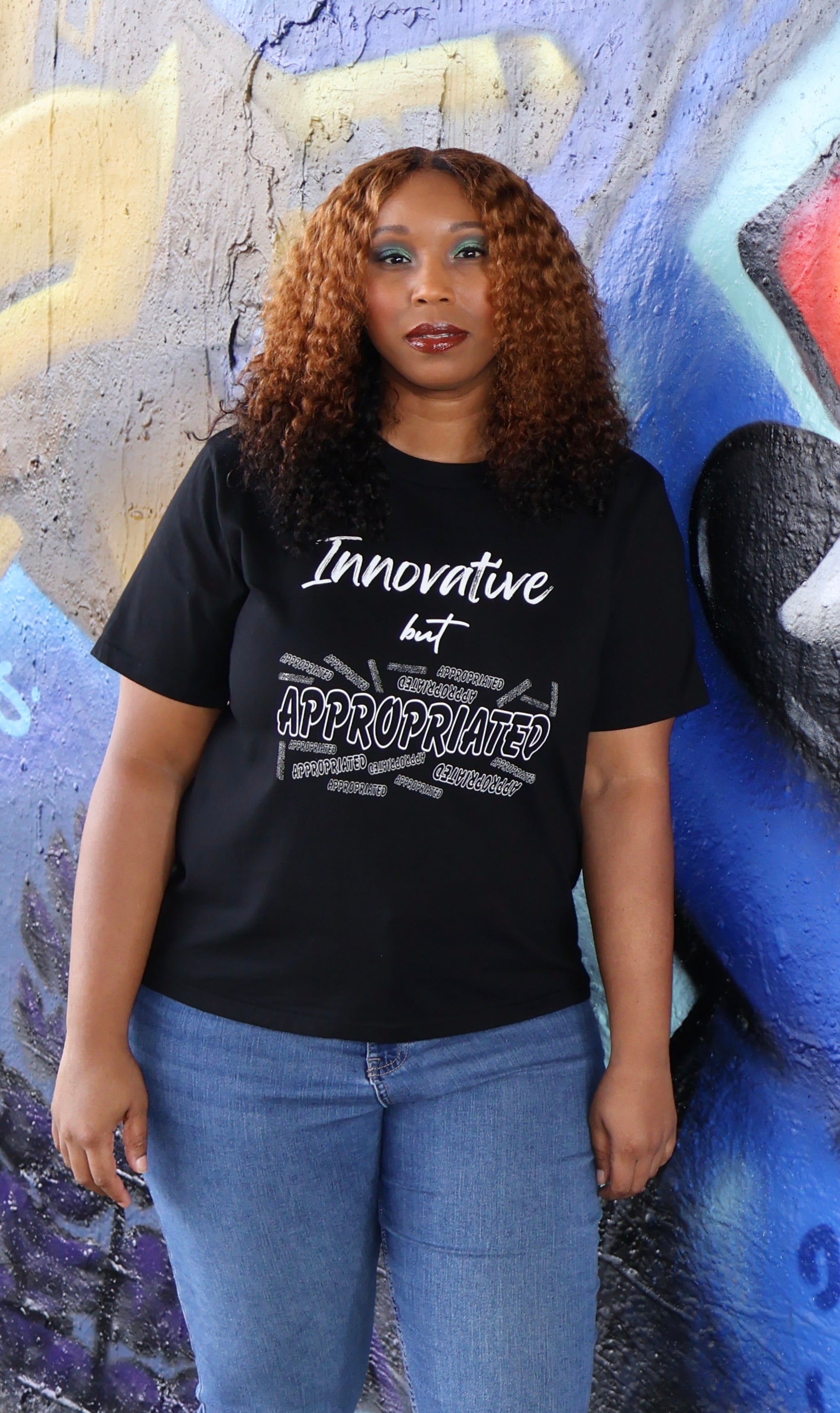 Woman wearing a black t-shirt with white 'Innovative but Appropriated' text and graphic design in front of a colorful mural.