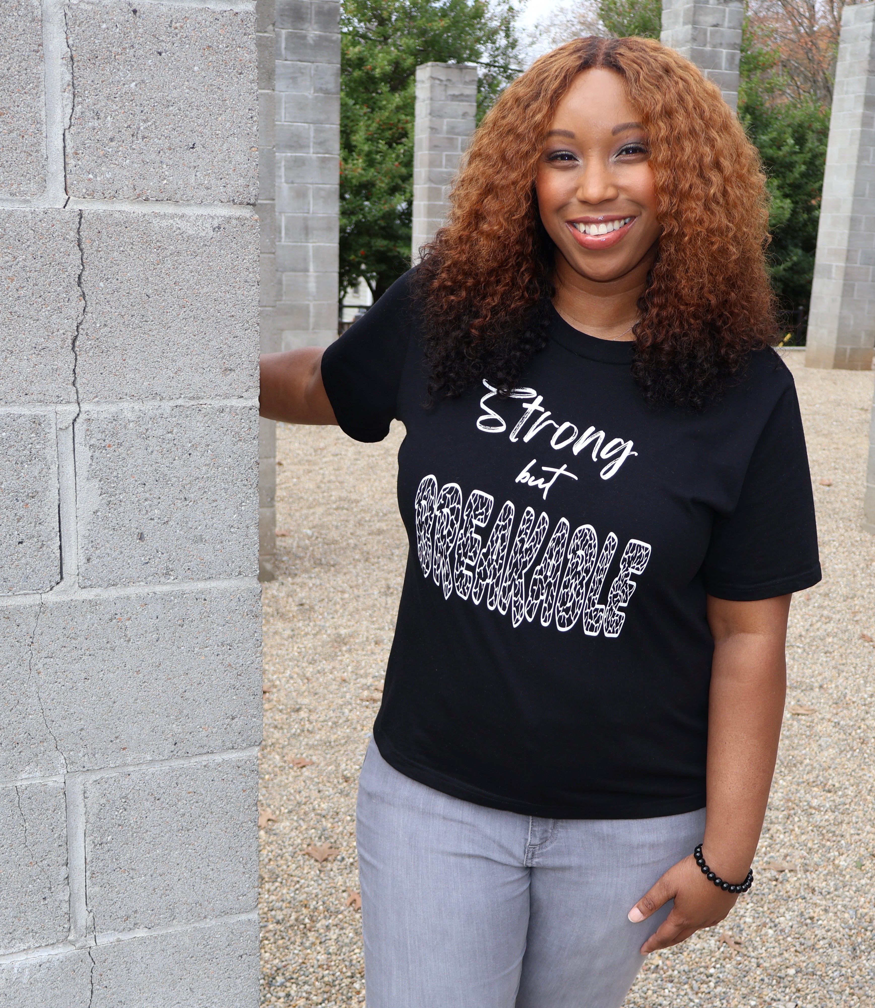 Woman wearing a white t-shirt with black 'Strong but Breakable' text and graphic design, standing outdoors next to a concrete column.