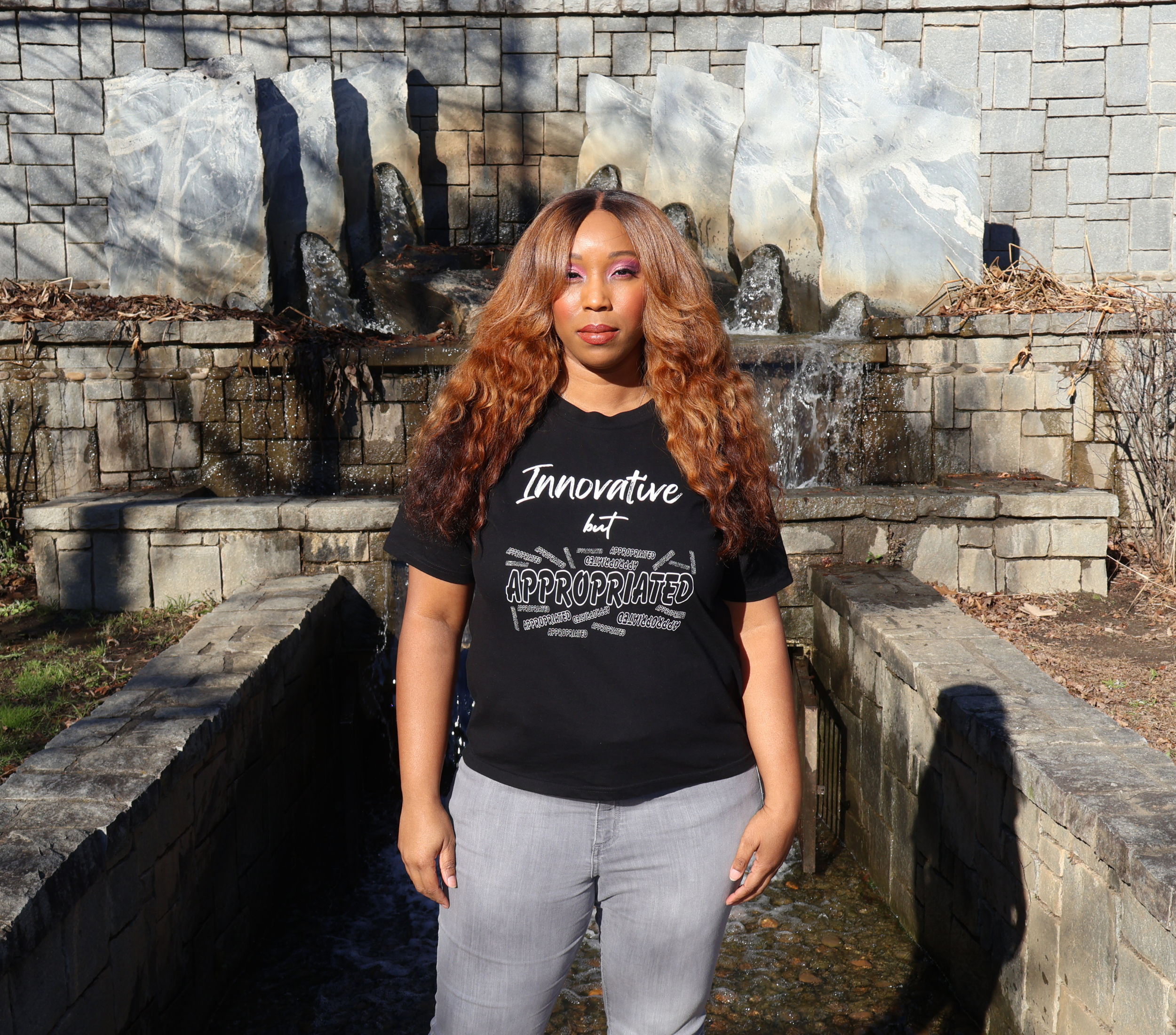 Woman wearing a black t-shirt with white 'Innovative but Appropriated' graphic design, standing in front of a stone wall waterfall.
