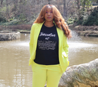 Woman wearing a bright yellow suit and black t-shirt with white 'Innovative but Appropriated' graphic design, standing next to a large tree log by a body of water.