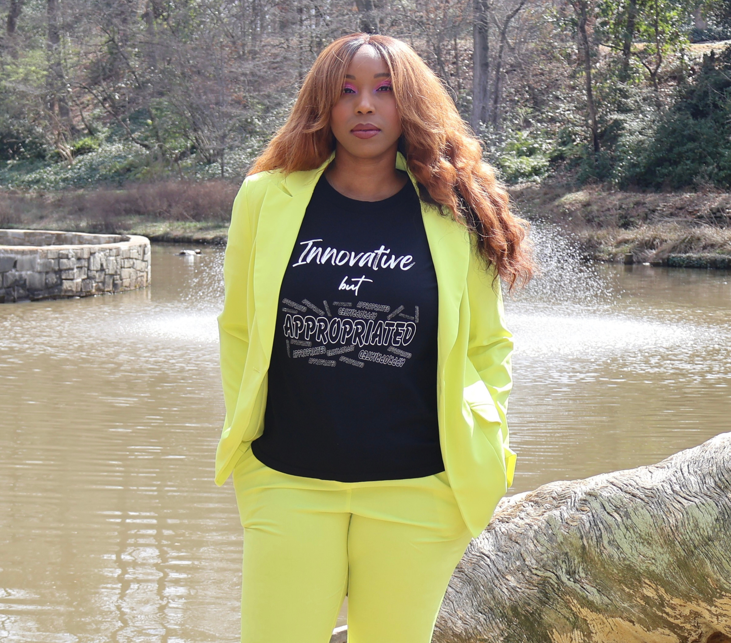 Woman wearing a bright yellow suit and black t-shirt with white 'Innovative but Appropriated' graphic design, standing next to a large tree log by a body of water.