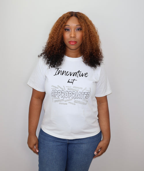 Woman wearing a white t-shirt with black 'Innovative but Appropriated' text and graphic design, standing against a gray wall