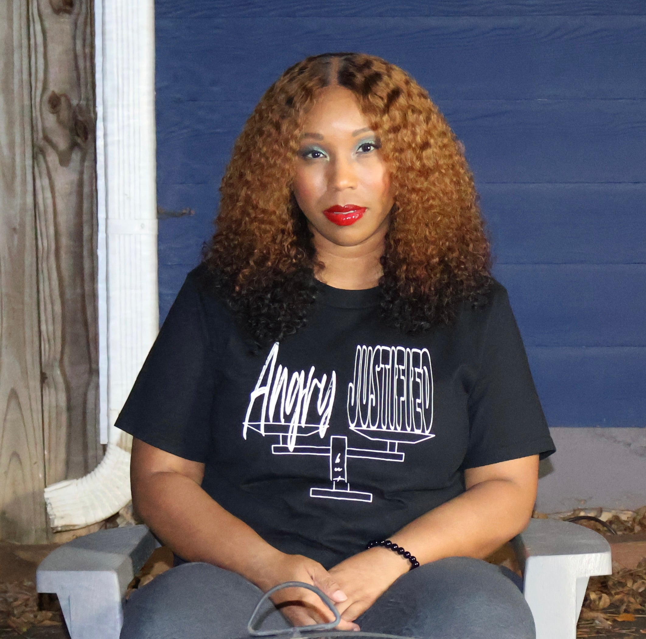 Woman wearing a black t-shirt with white 'Angry but Justified' text and graphic design, sitting on a grey chair against a blue wall.