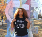 Woman wearing a black t-shirt with white 'Resilient but Tired' graphic design, standing in front of a 'Black Lives Matter' mural.