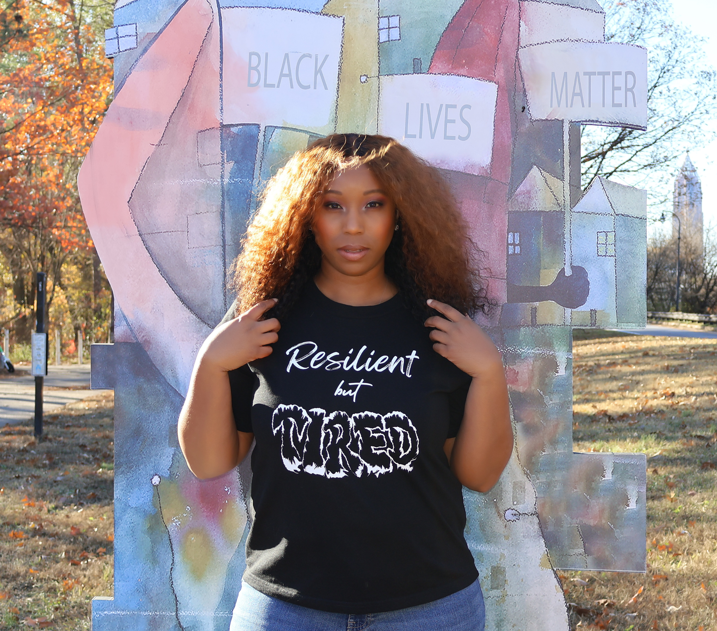Woman wearing a black t-shirt with white 'Resilient but Tired' graphic design, standing in front of a 'Black Lives Matter' mural.