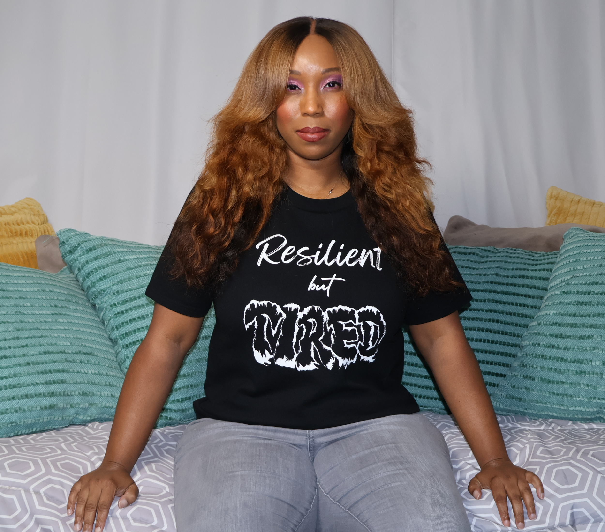 Woman sitting on a couch with green, yellow, and gray pillows, wearing a black t-shirt with white 'Resilient but Tired' graphic design.