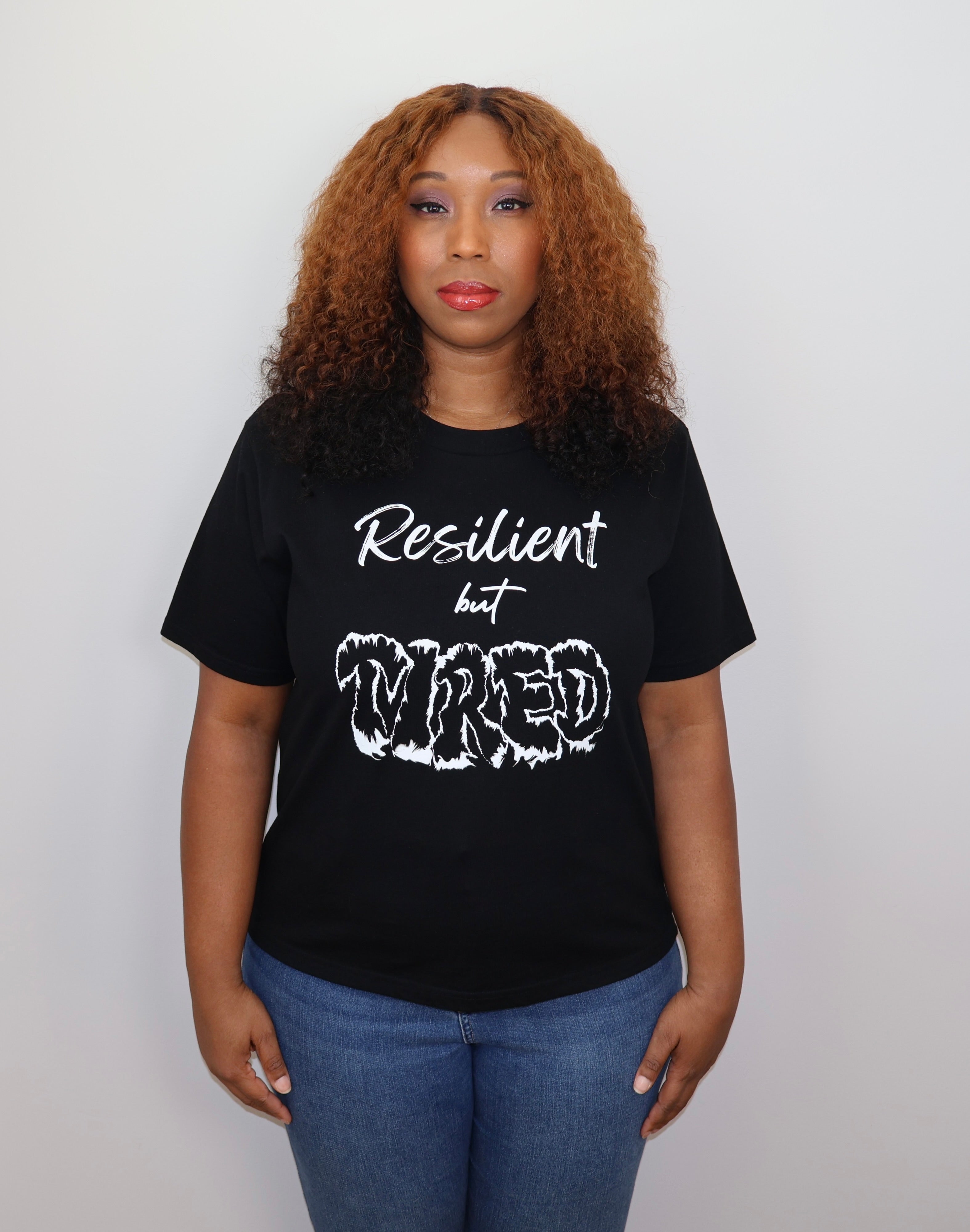 Woman wearing a black t-shirt with white 'Resilient but Tired' text and graphic design, standing against a gray wall