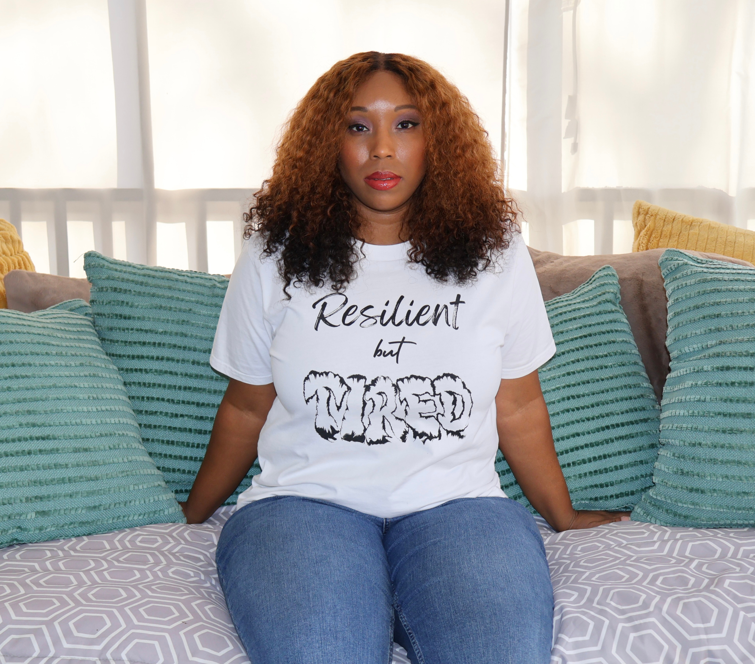 Woman sitting on a couch with green, yellow, and gray pillows, wearing a white t-shirt with black 'Resilient but Tired' graphic design.