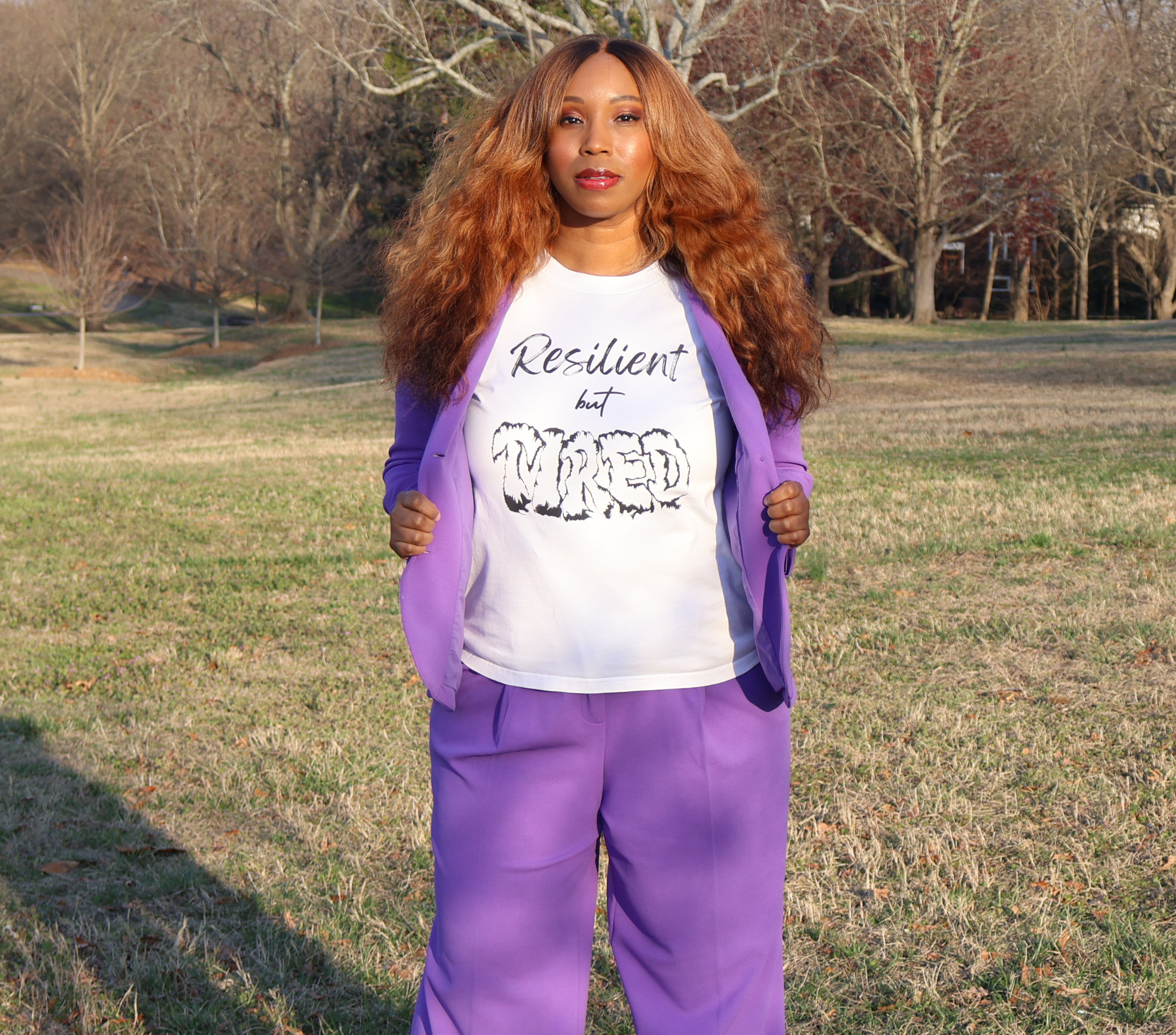 Woman wearing a purple suit with a white t-shirt with black 'Resilient but Tired' graphic design standing outside in an open field with grass and trees.