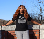 Woman wearing a black t-shirt with white 'Strong but Breakable' graphic design, standing against a brick wall with trees in the background.