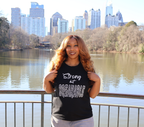 Woman wearing a black t-shirt with white 'Strong but Breakable' graphic design, standing by a lake with trees and city buildings in the background.