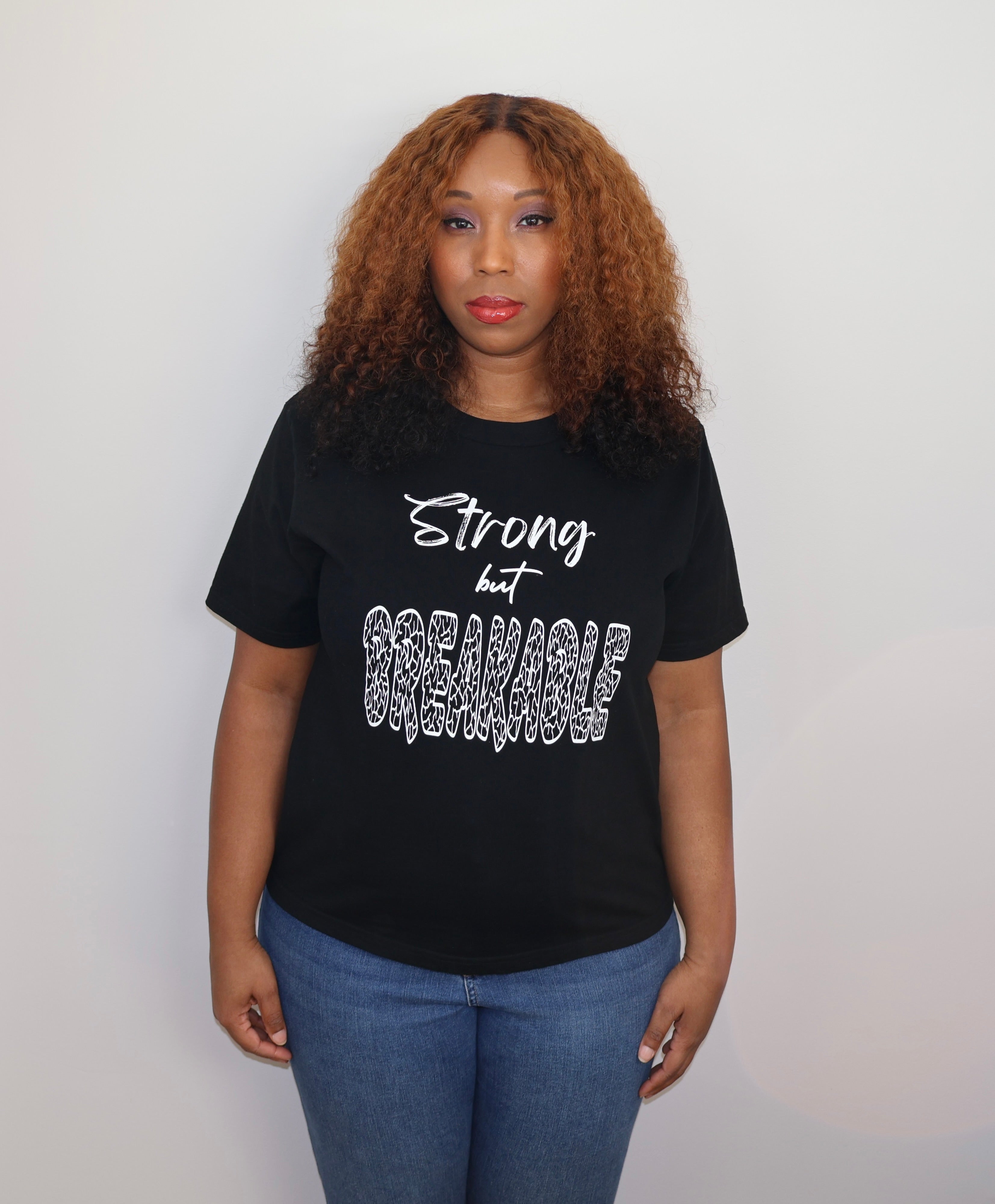 Woman wearing a black t-shirt with white 'Strong but Breakable' text and graphic design, standing against a gray wall