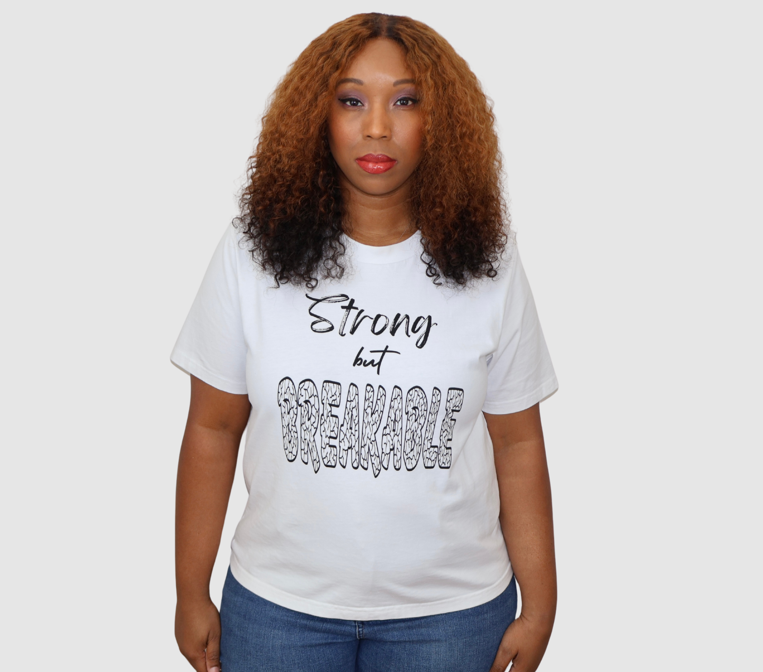 Woman wearing a white t-shirt with black 'Strong but Breakable' graphic design, standing against a gray background.