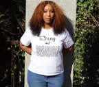 Woman wearing a white t-shirt with black 'Strong but Breakable' graphic design, standing outside against a concrete pillar with trees.