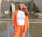Woman wearing an orange suit with a white t-shirt with black 'Strong but Breakable' graphic design, standing in front of a stone wall waterfall.
