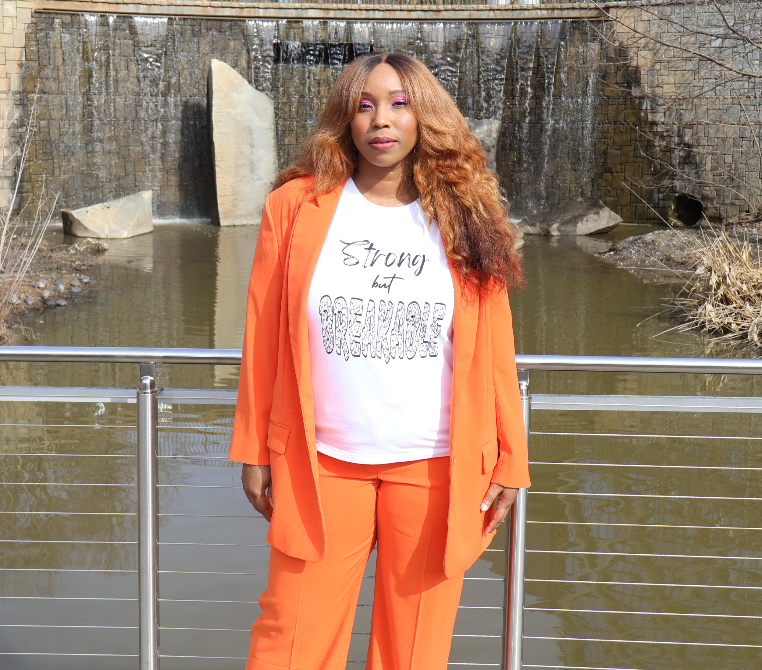 Woman wearing an orange suit with a white t-shirt with black 'Strong but Breakable' graphic design, standing in front of a stone wall waterfall.